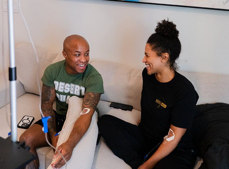 Two people sitting on a couch, receiving IV drips, smiling and talking. IV stand in view.