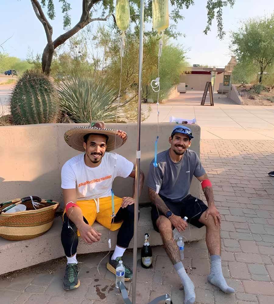 Two men in athletic and festive attire sitting outdoors by a cactus, receiving post-activity IV hydration for recovery.