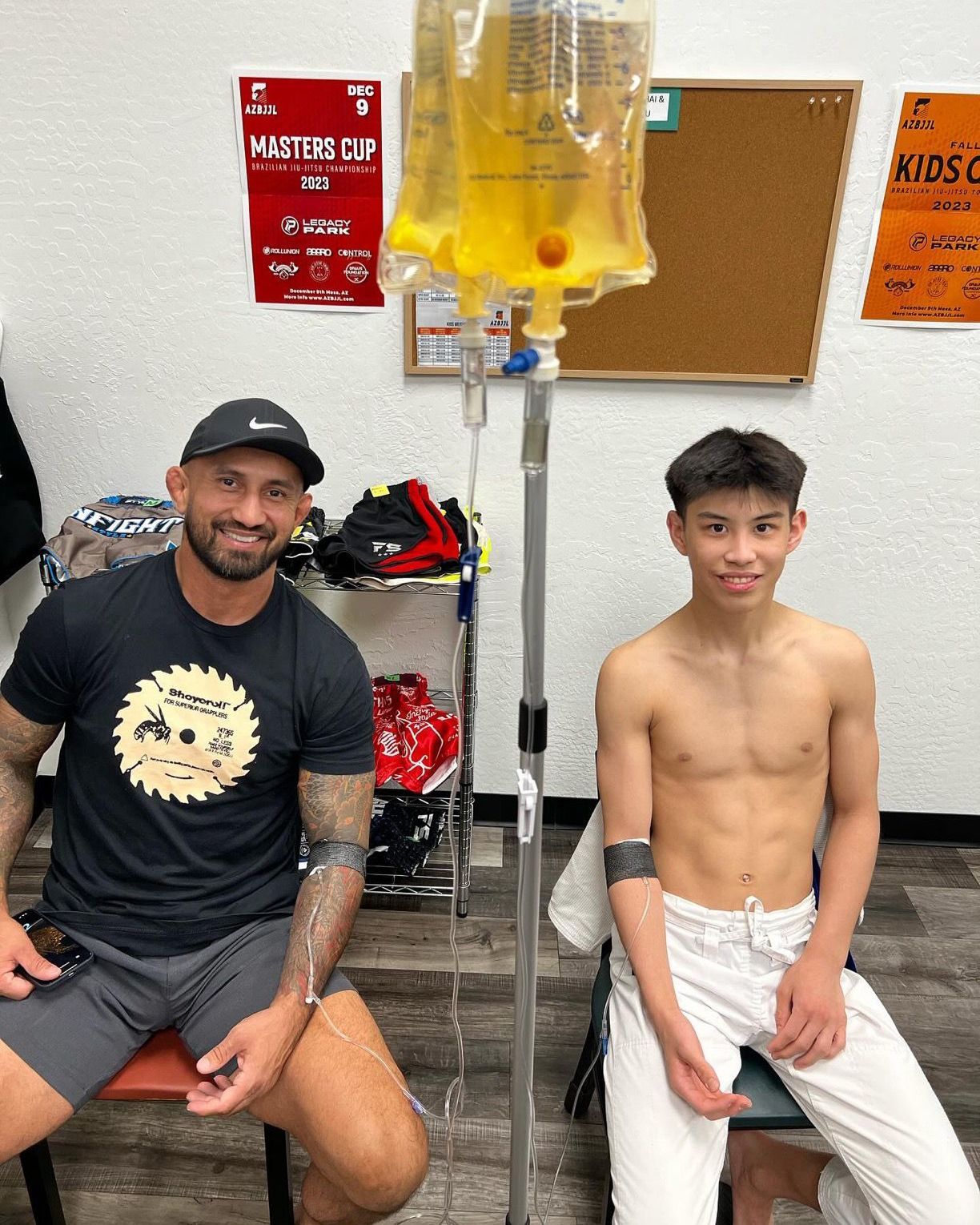 A smiling coach and a young athlete sit in a gym while receiving intravenous hydration therapy from a hanging IV bag.