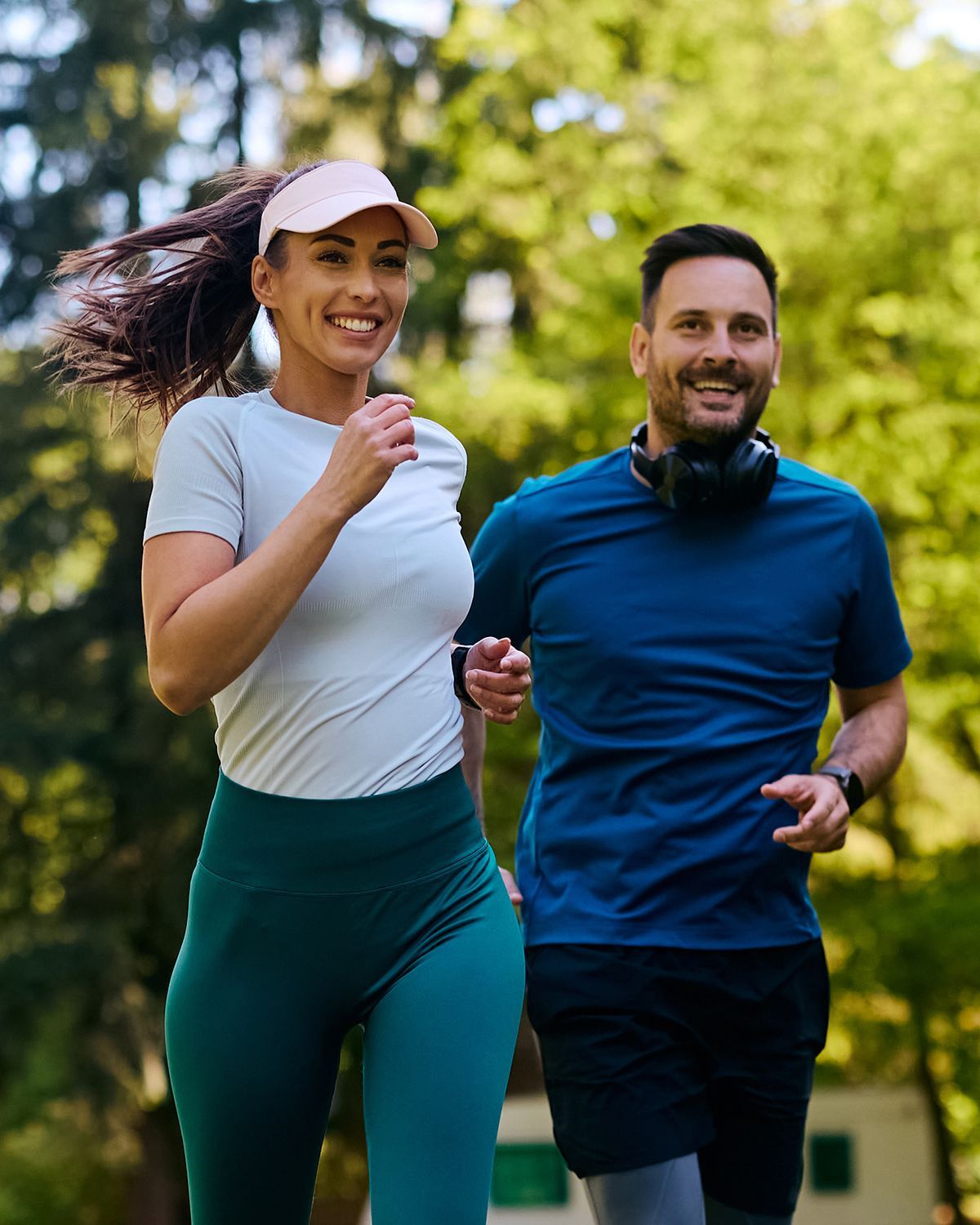 A smiling couple jogs together through a sunlit, wooded park, both dressed in activewear.
