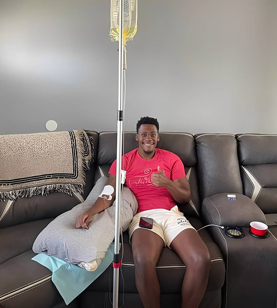 A smiling man in athletic shorts gives a thumbs-up while receiving a mobile IV hydration drip infusion on his couch for fast sports recovery.