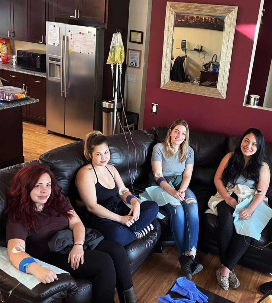 Four people receive IV infusions while sitting on a couch in a living room.