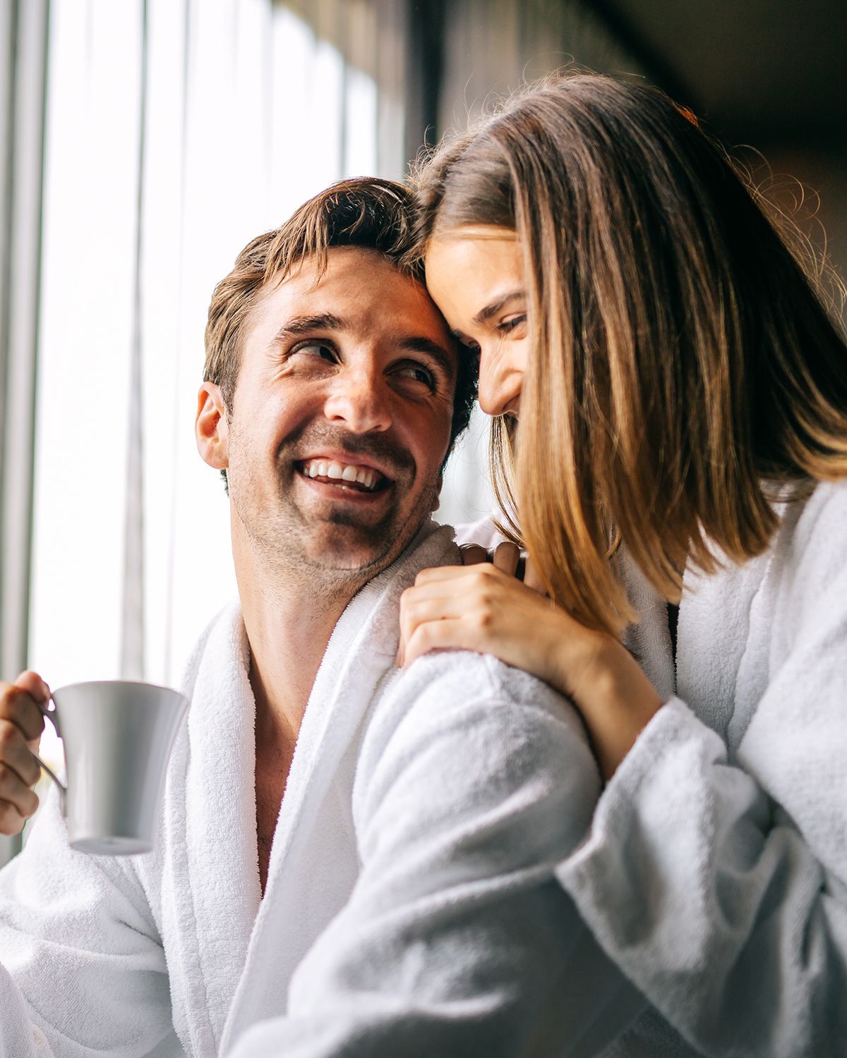A couple wearing white bathrobes smiling at each other while indoors, one holding a white mug.