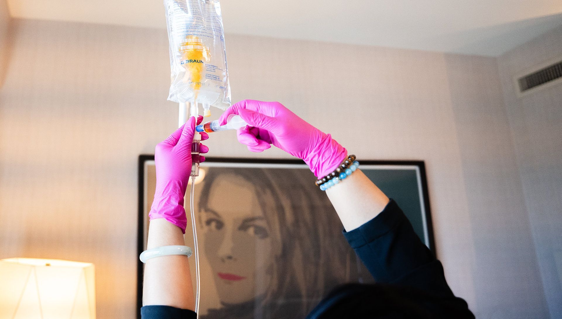 Person in pink gloves adjusts IV bag hanging in a room.