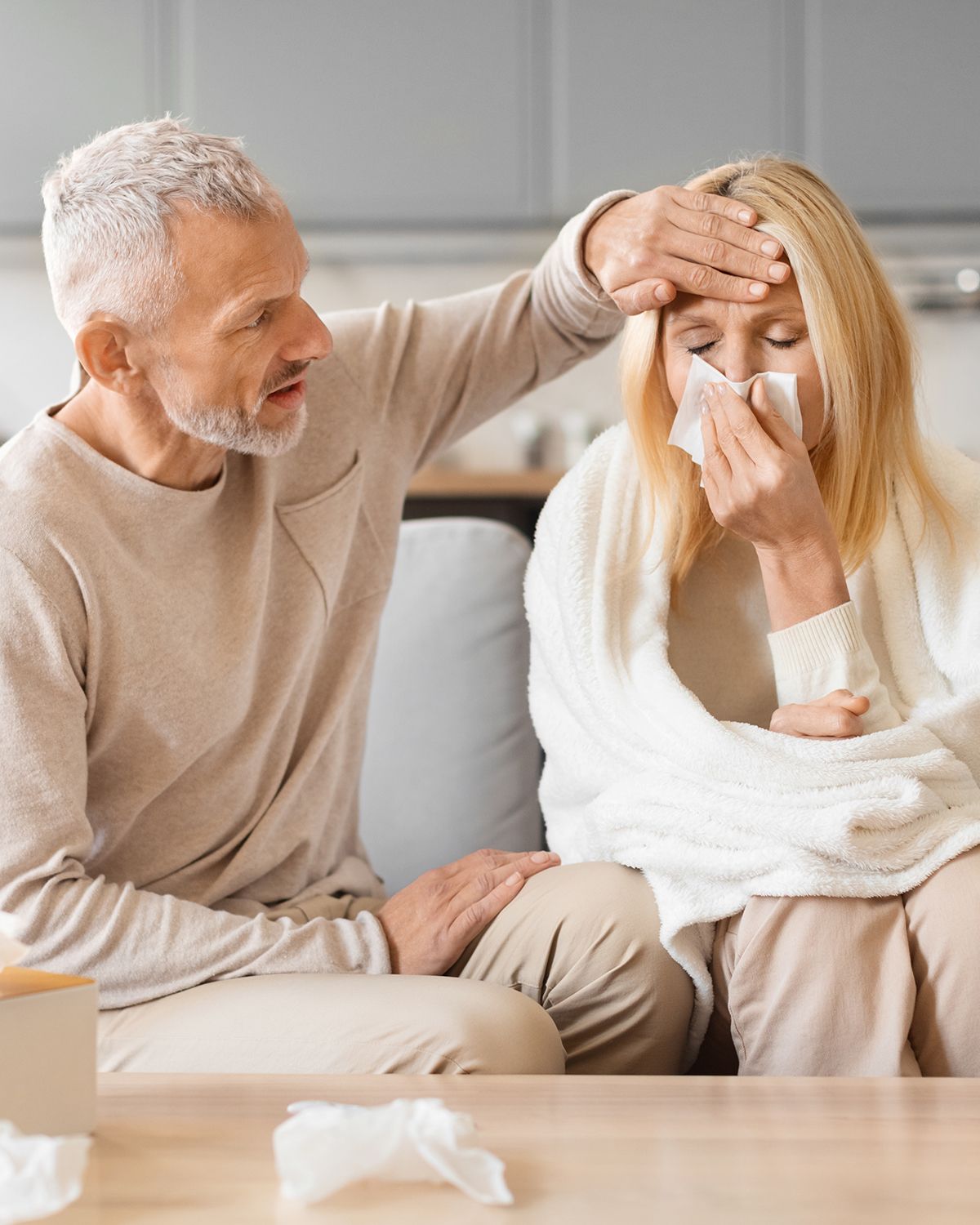 A person places a hand on another's forehead to check for a fever as they hold a tissue to their nose while wrapped in a blanket.