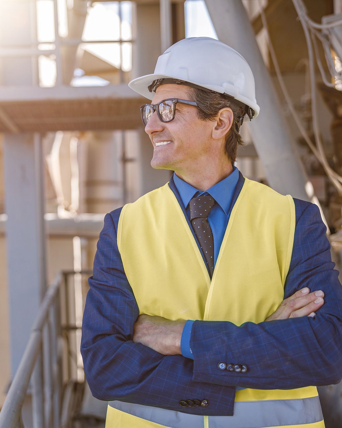 A professional wearing a hard hat, suit, and safety vest stands at an industrial site with arms crossed, smiling.