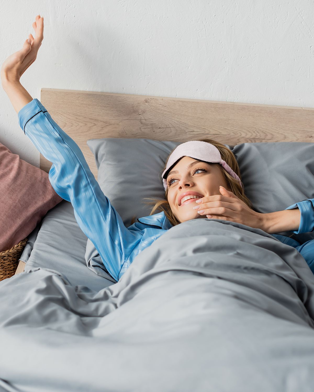 A person wearing a sleep mask and blue pajamas wakes up in bed, smiling and reaching upward.