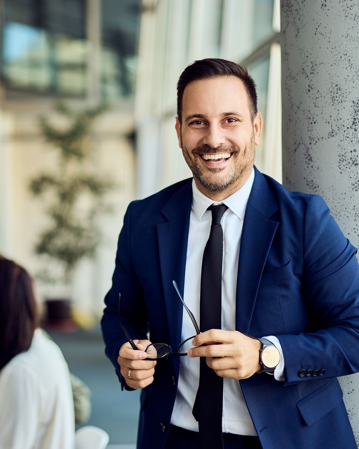 A smiling professional in a navy suit and tie holds glasses, standing in a brightly lit office setting.