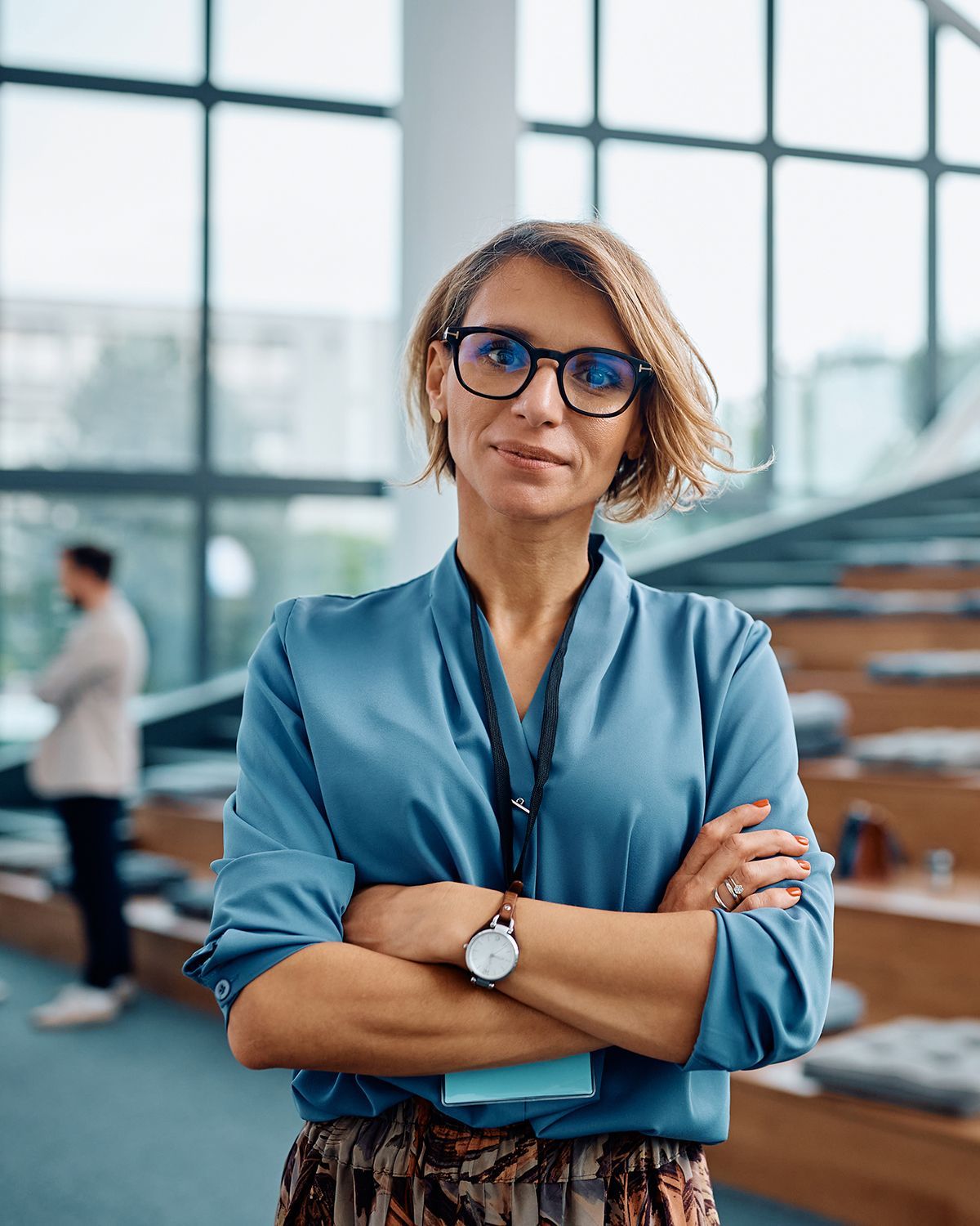 A person with short hair and glasses wearing a blue blouse and watch stands with arms crossed in a bright, modern office.