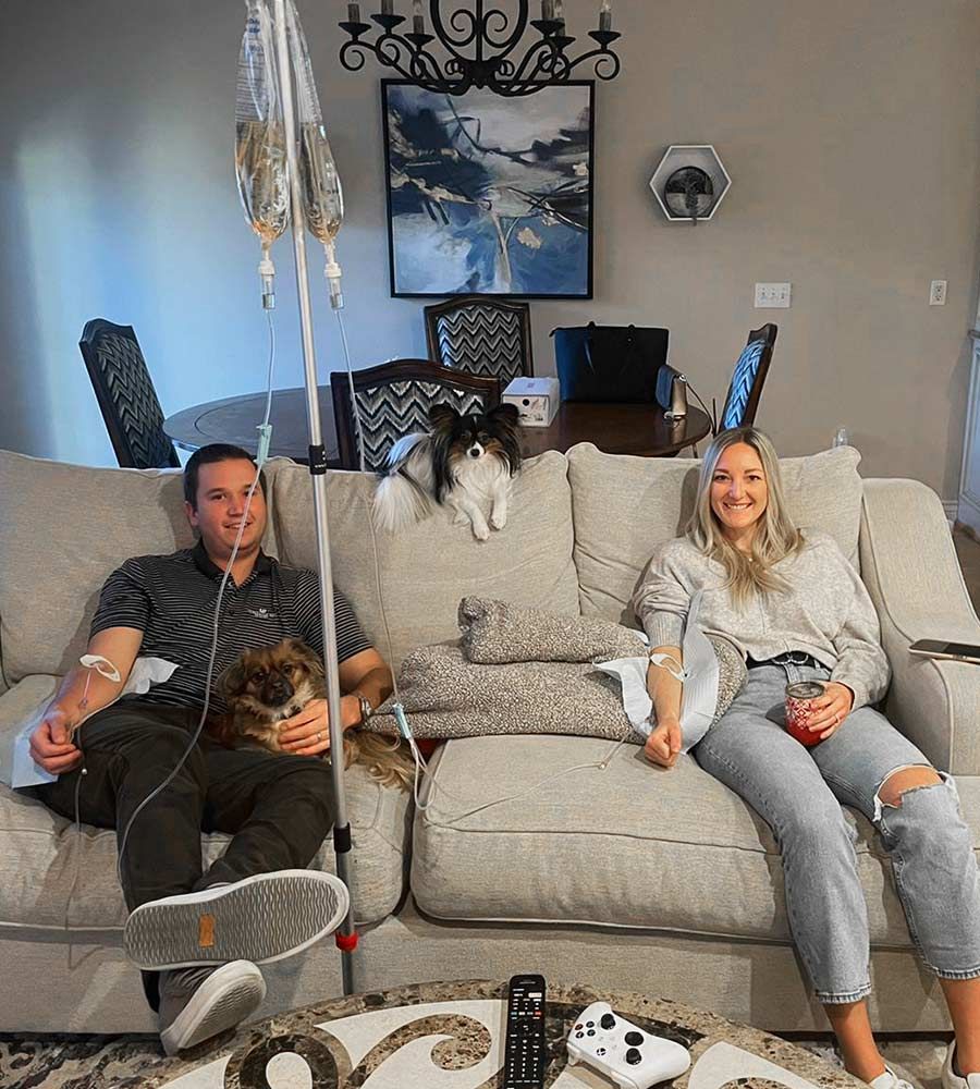 Smiling couple relaxing on a couch with two small dogs, both receiving an IV hydration drip at home.