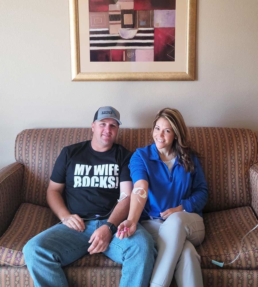 A happy couple sits together on a couch, both receiving a mobile IV drip infusion for couples' wellness or recovery.