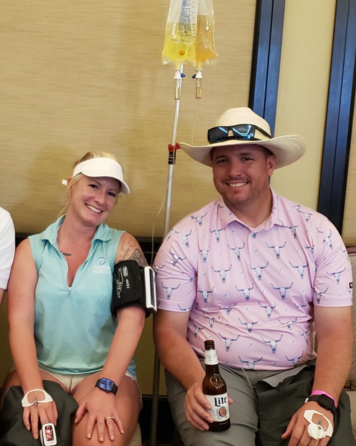 Two people wearing medical IVs and monitors on their arms, sitting together while one holds a bottle of beer.