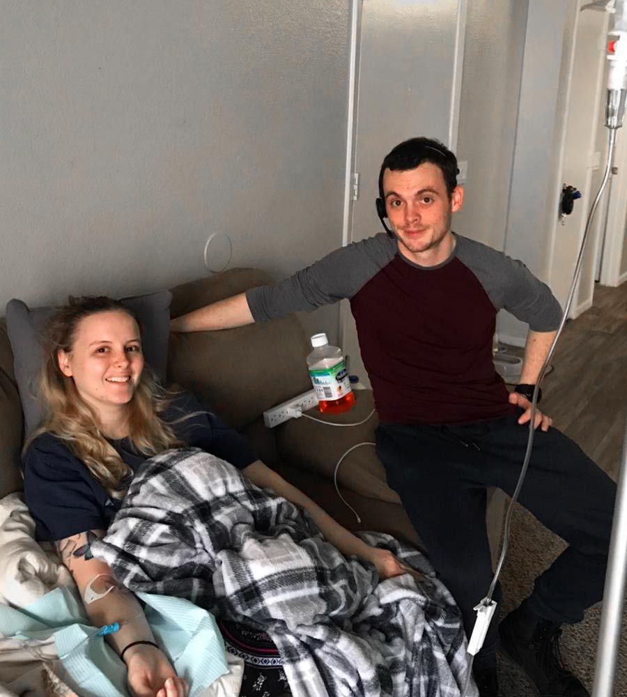 Woman receiving IV in bed, smiling; man stands beside her, smiling, with headset.