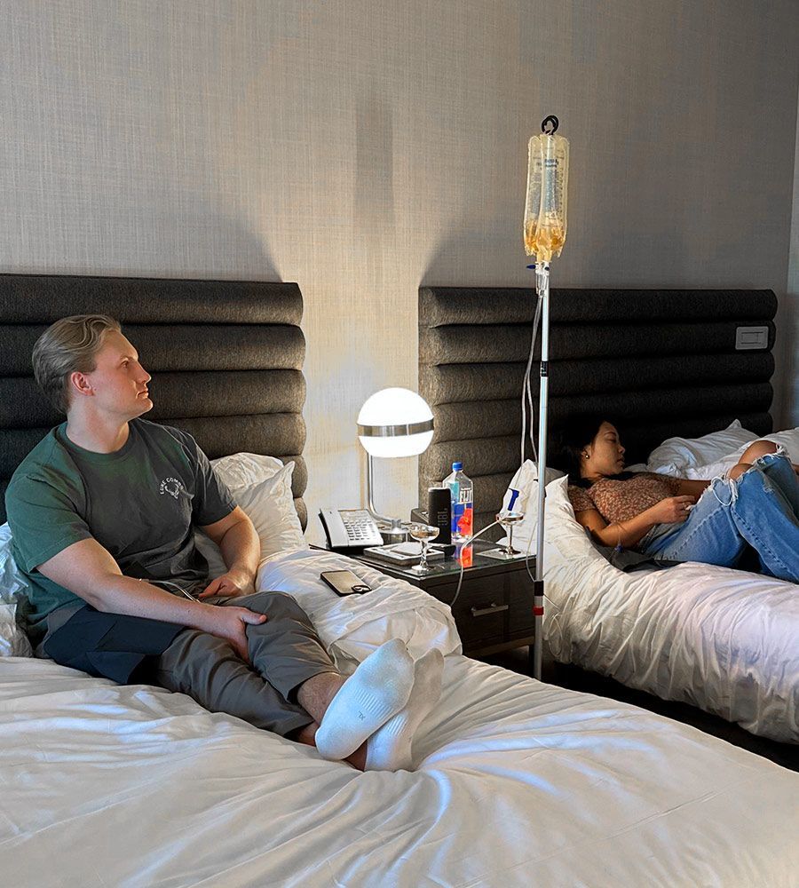 Male and female clients relaxing on separate beds in a dual-bed hotel room while receiving mobile IV hydration therapy for travel recovery, showcasing convenient, in-room service for companions.