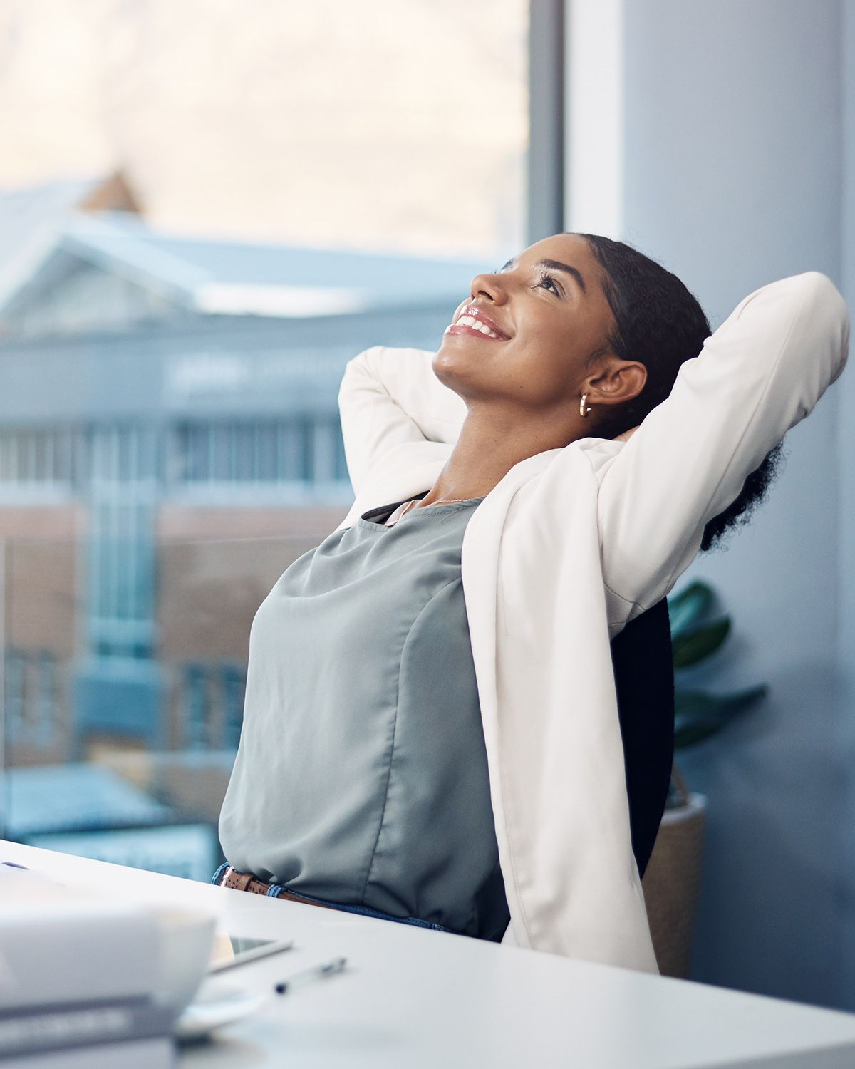 A person in business attire leans back in an office chair with hands behind their head, smiling while looking upward.