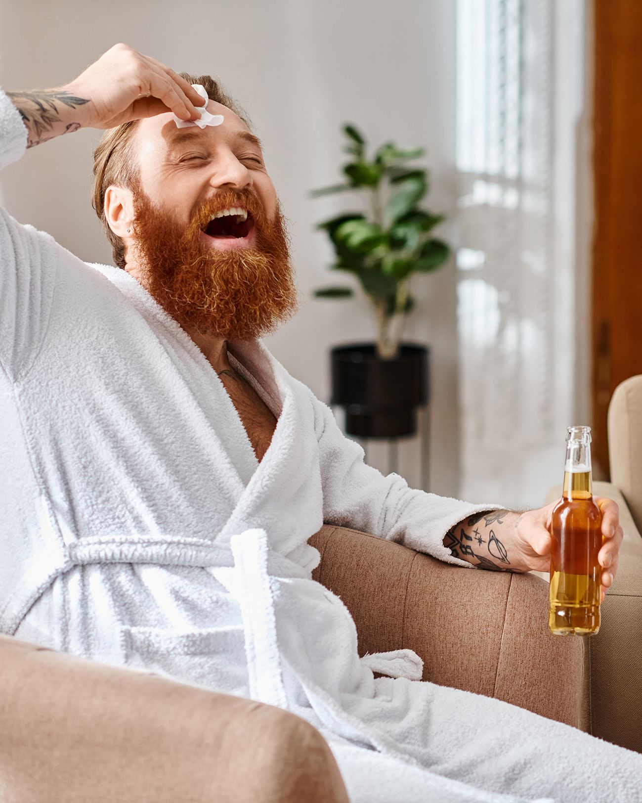 A man with a ginger beard wearing a white bathrobe laughs while applying a patch to his forehead, holding a beer bottle.
