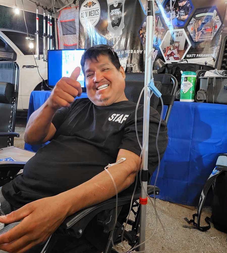 Man in chair with IV, giving thumbs up. Indoors with blue table and banners.