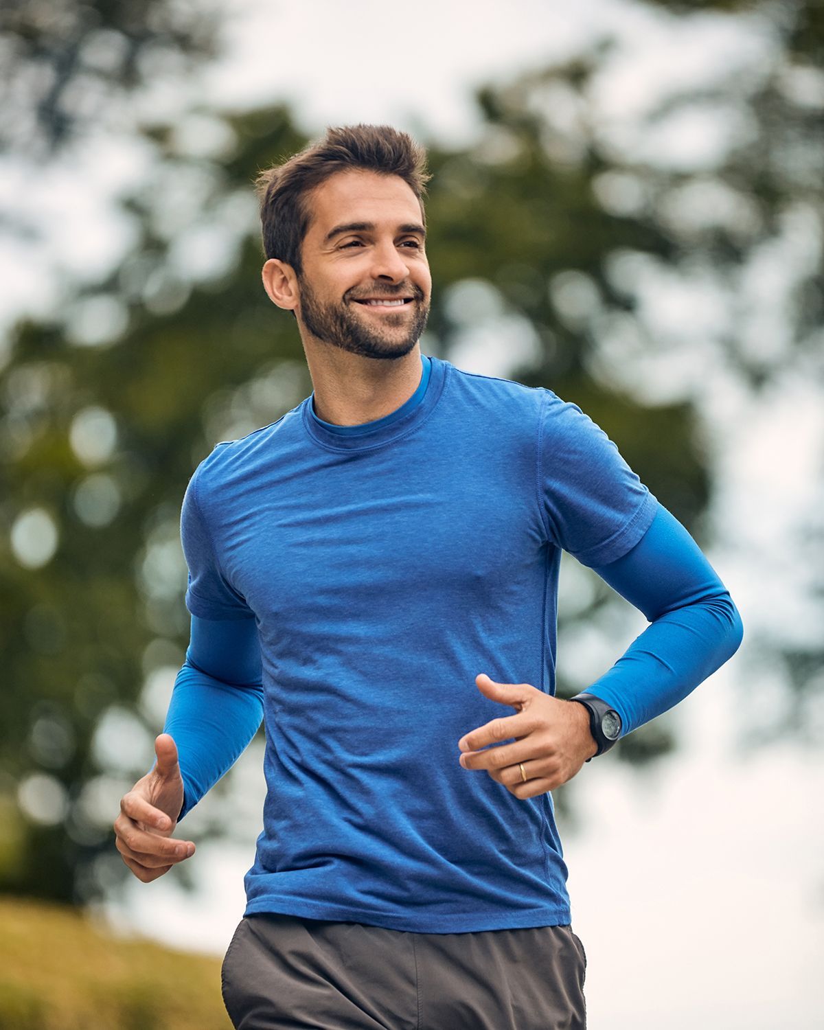 A person smiling while jogging outdoors, wearing a blue long-sleeved shirt, dark shorts, and a smartwatch.