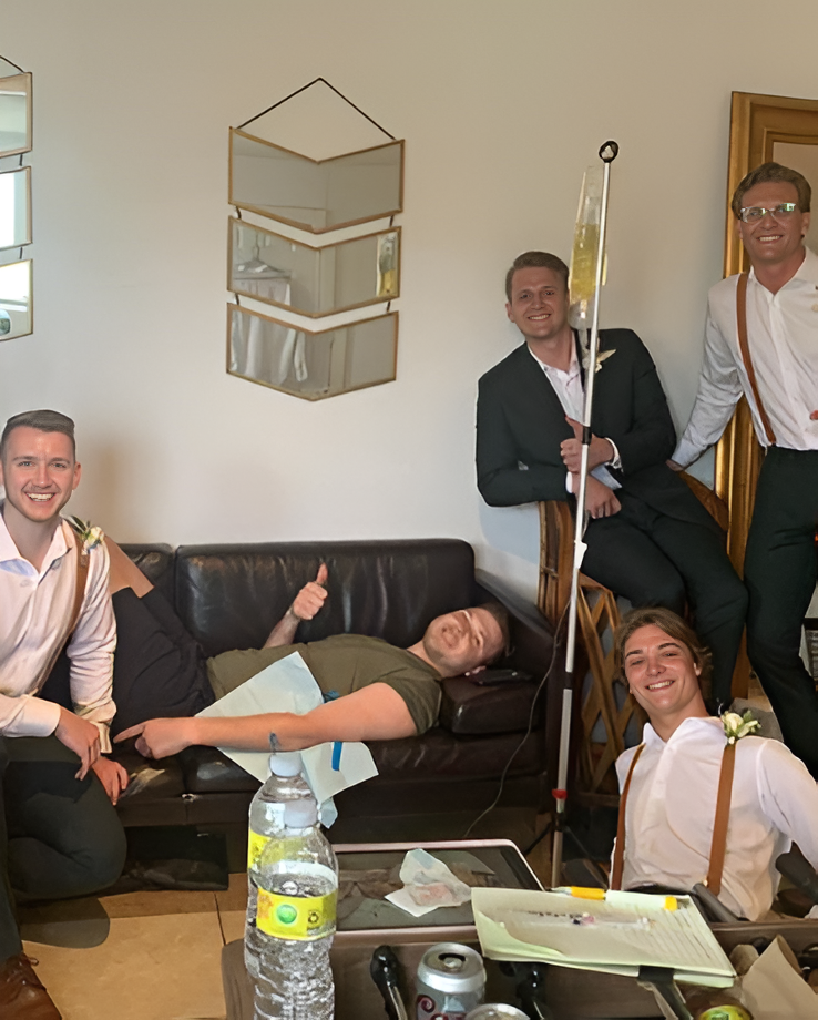 Five people in formal attire and casual clothes gather in a room, with one person lying on a sofa hooked to an IV stand.