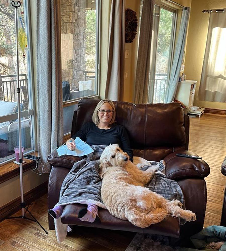 Smiling woman receiving mobile IV vitamin therapy in a brown leather recliner at home, relaxing under a blanket with a large therapy dog companion.