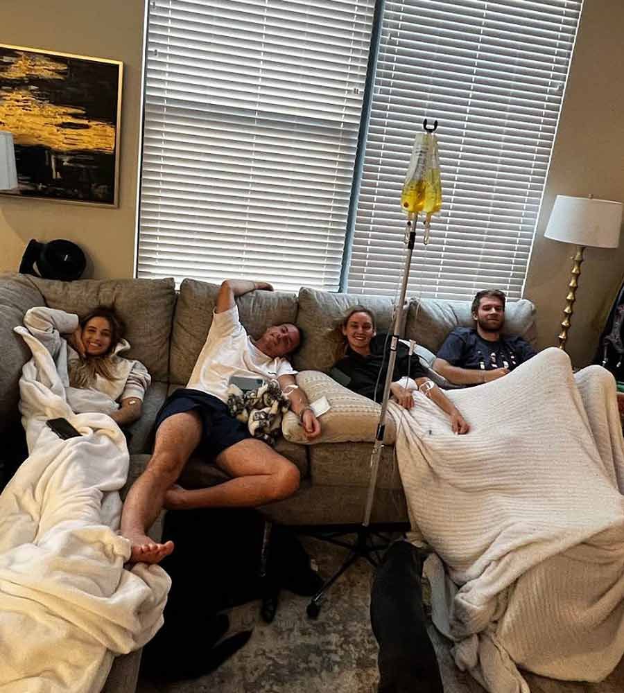 Four young adults relaxing on a couch under blankets, receiving a group IV hydration drip in a living room setting.