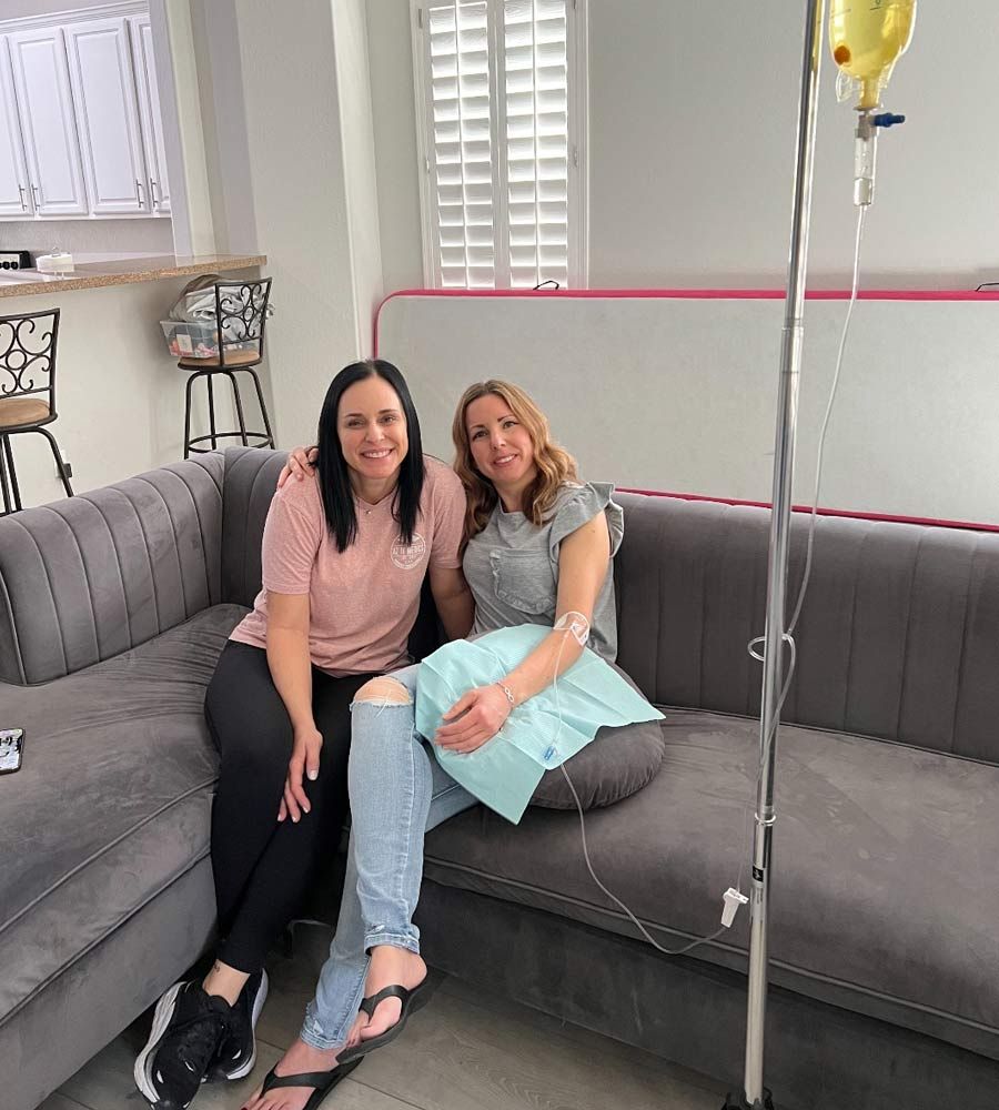 Two women on a couch, one receiving IV fluids. Living room setting with medical equipment present.