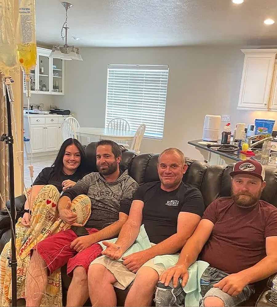 Four adults sitting on a dark leather sofa in a home with a kitchen visible in the background, receiving IV infusions.