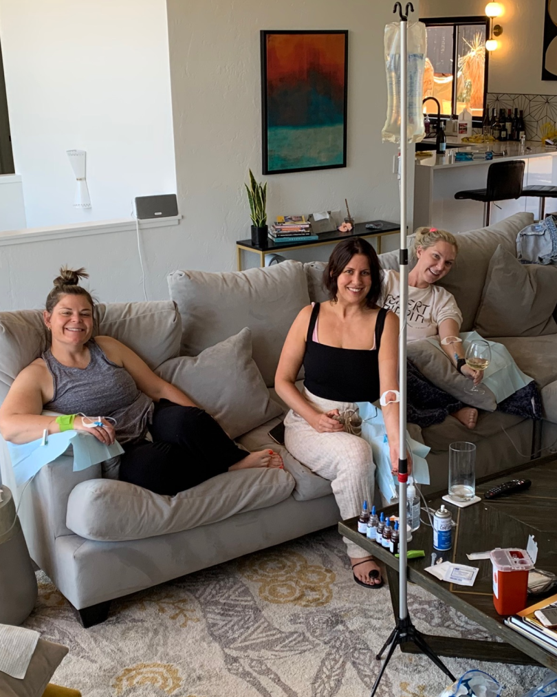 Three people sit on a couch in a living room, each receiving an intravenous infusion from a shared medical stand.