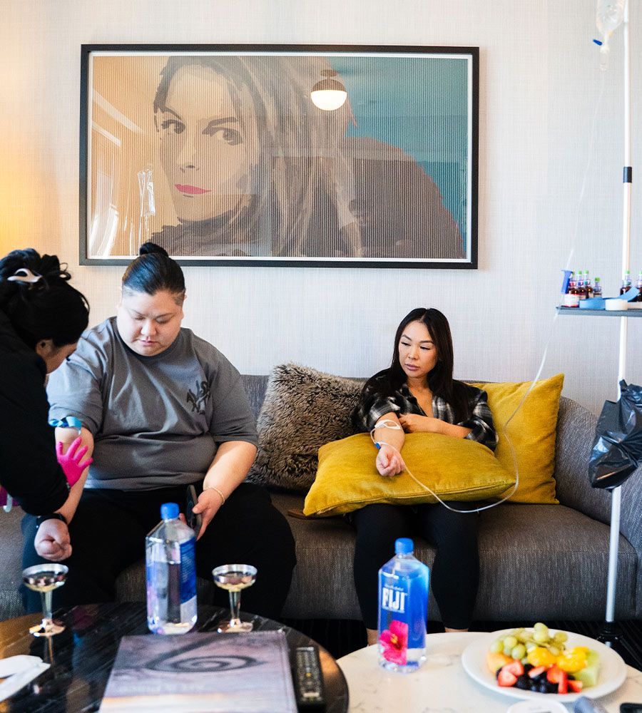 Two women receiving simultaneous mobile IV hydration therapy from a nurse on a stylish couch in a hotel suite, ideal for travel recovery, groups, or bachelor/bachelorette parties.