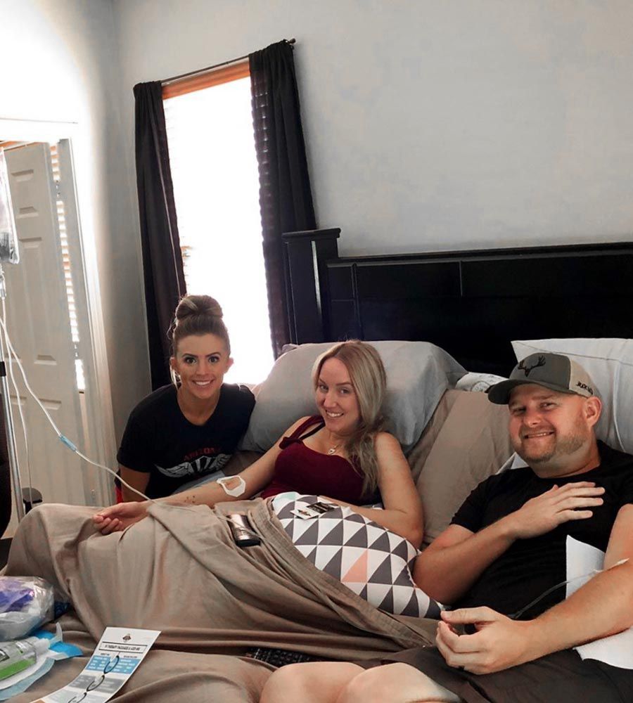 Three people sit on a bed with IV lines. One woman smiles, another woman and a man look at the camera. Neutral background.