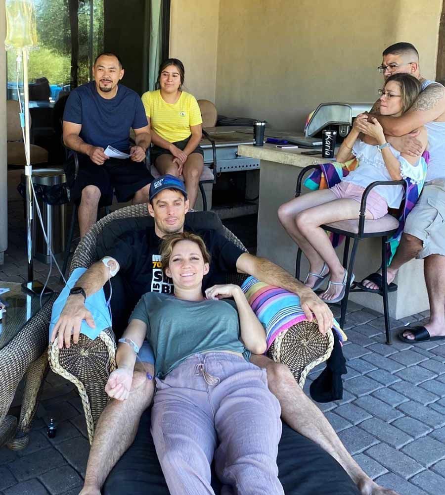 Group of people outdoors: one couple seated, others standing/sitting nearby. Neutral expressions, patio setting.