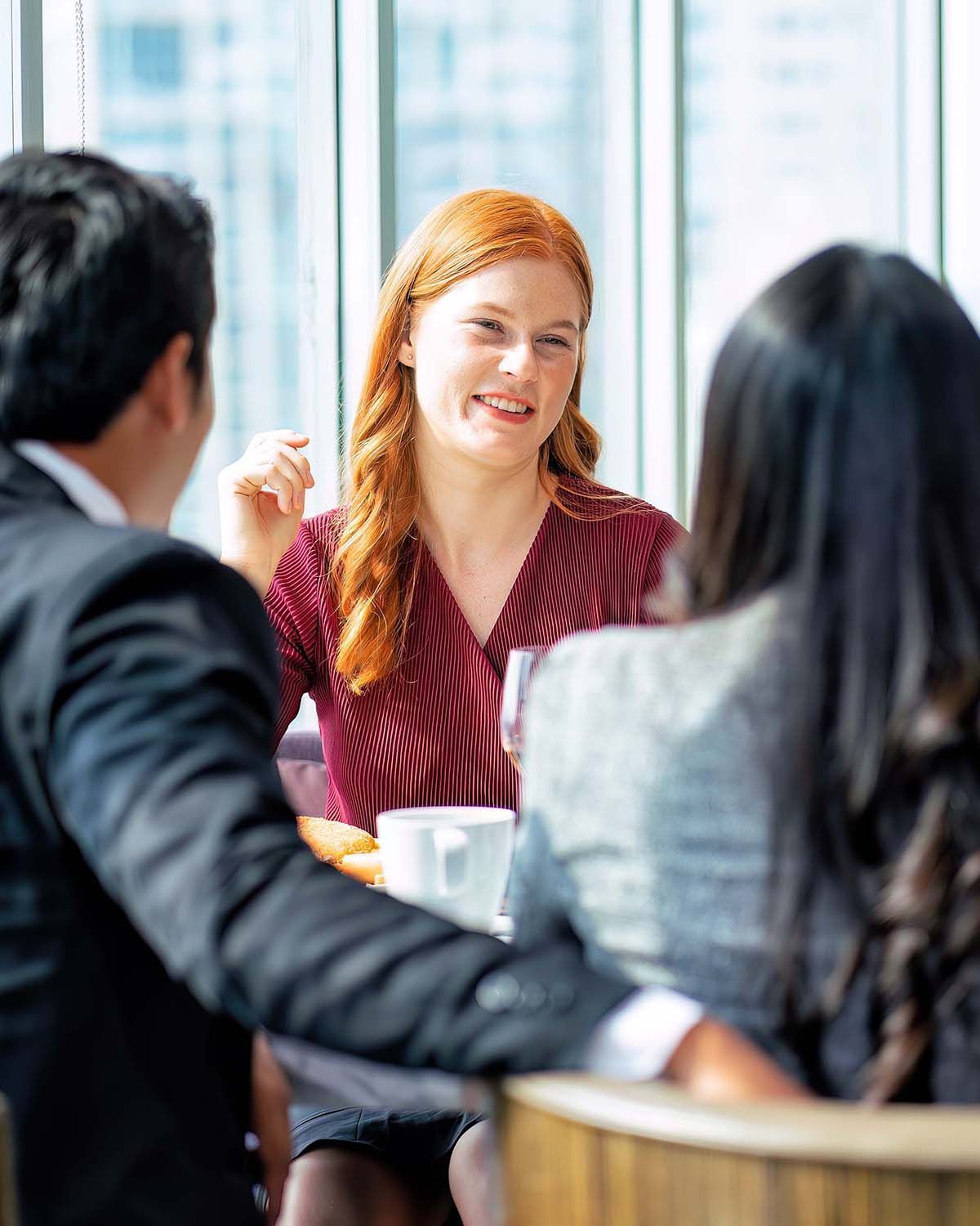 Three people in professional attire having a conversation at a table in a brightly lit office setting with large windows.