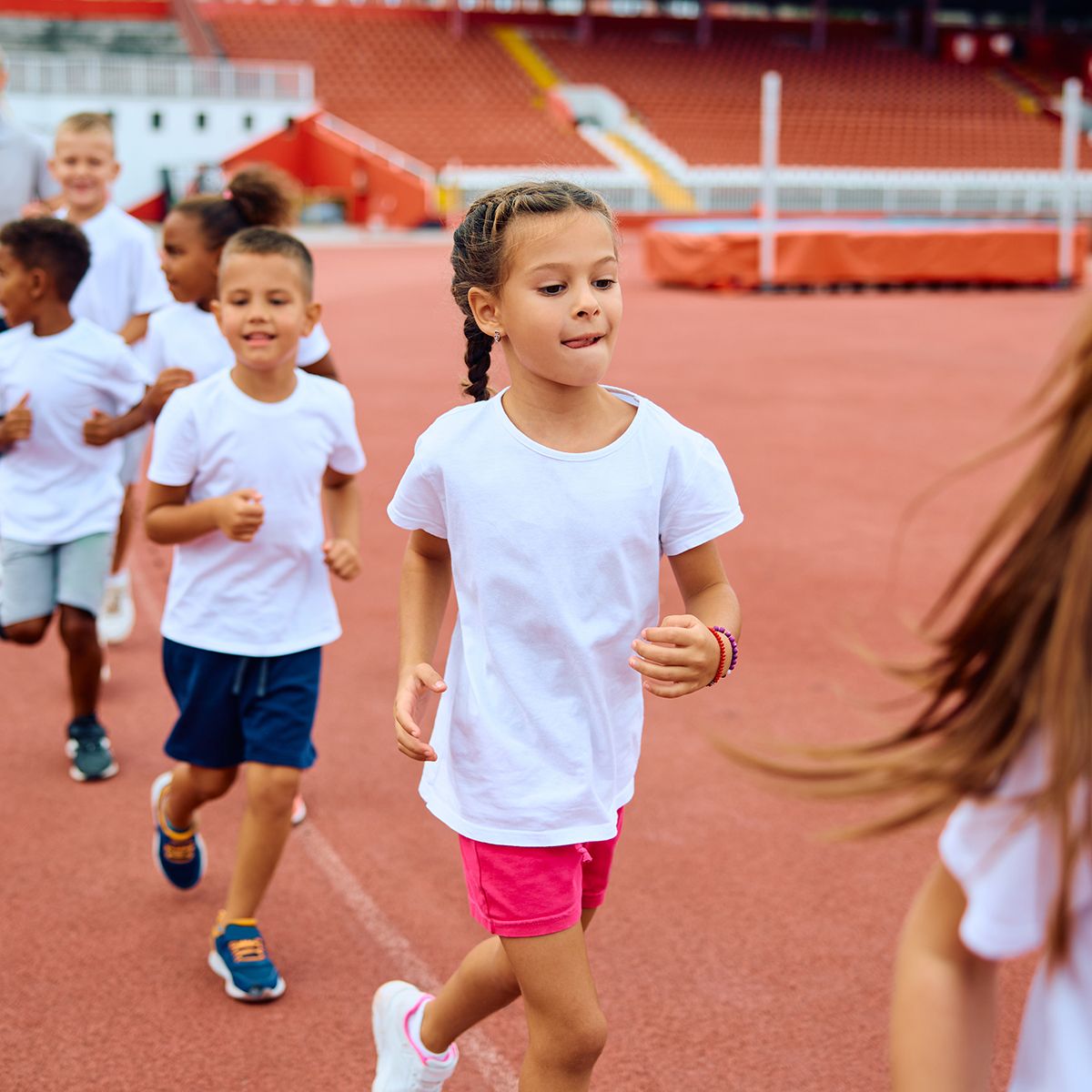 A group of children in white shirts jogging on an orange track at a sports stadium.