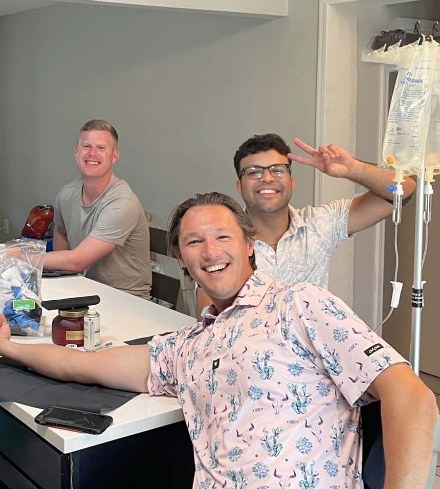 Three smiling men receiving group IV hydration or vitamin therapy in a casual setting with an IV pole and bags visible.