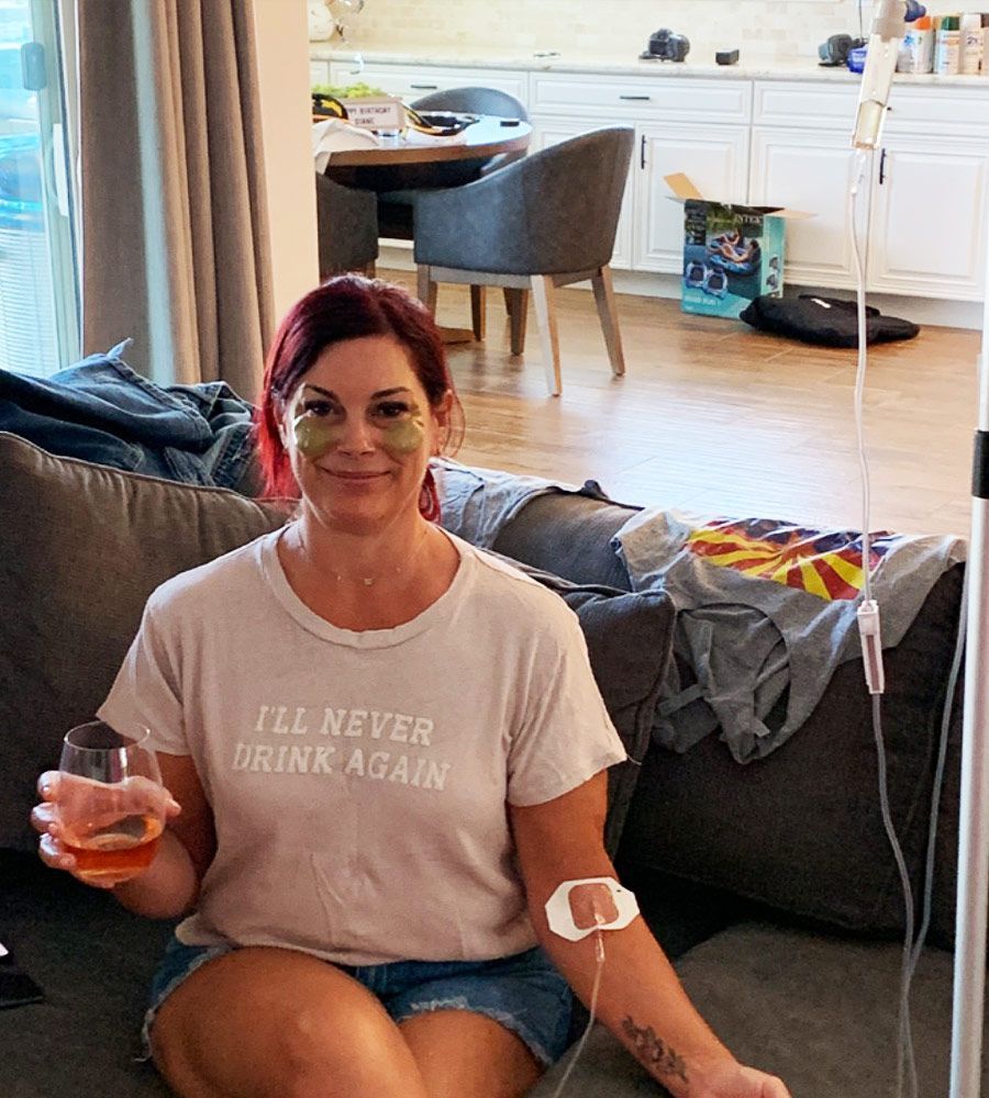 Woman on a couch with IV drip, eye patches, and drink; wearing a shirt that says 