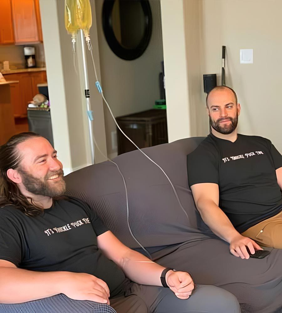 Two men relaxing on a sofa, one receiving a mobile IV hydration drip for quick recovery and energy, highlighting convenient group wellness service.