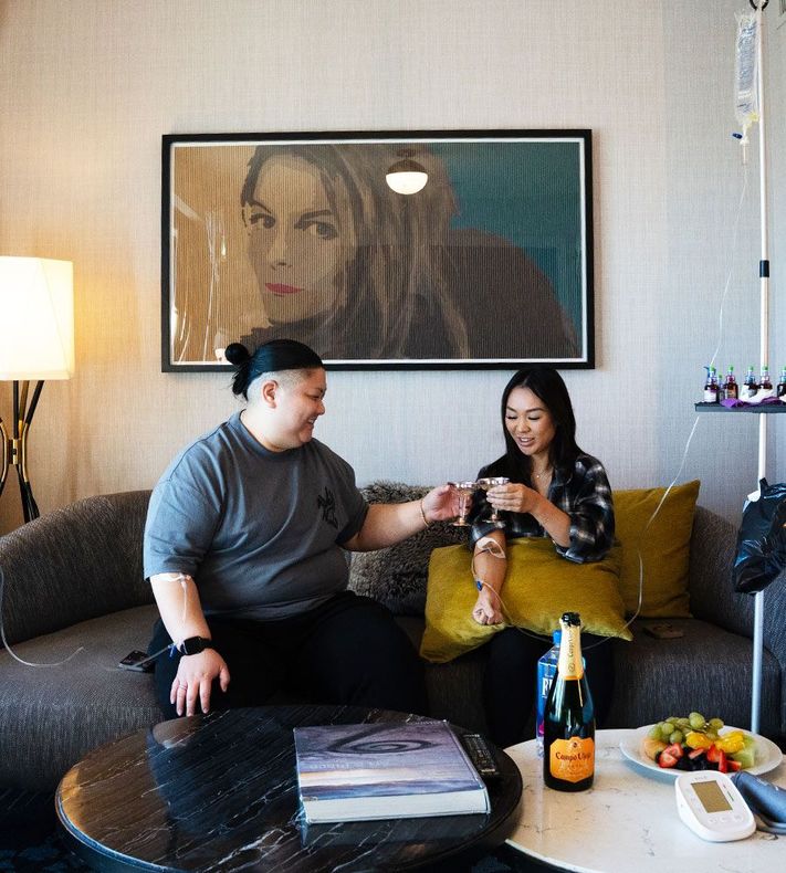 Two people in a room; one receiving an IV drip, the other holding medication, seated on a couch with champagne and fruit.