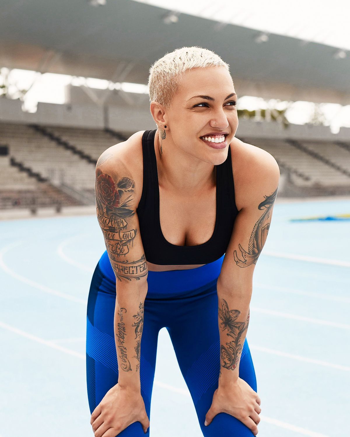 A person with short blonde hair and tattoos smiles while resting with hands on knees on a blue outdoor running track.