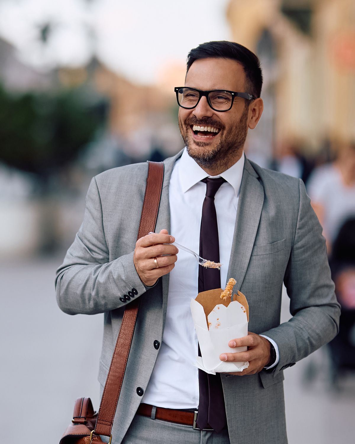 A smiling professional wearing a grey suit and glasses eats from a takeout container while walking outdoors.