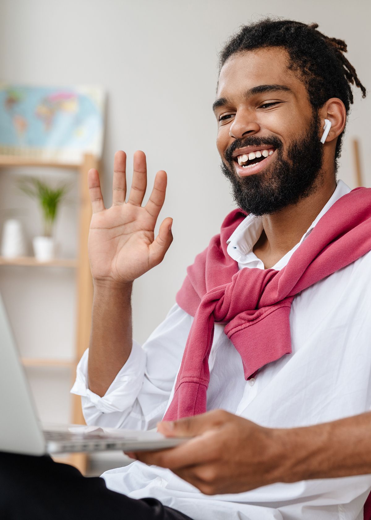 A person with dreadlocks and a beard smiles and waves while on a video call, wearing a white shirt and a pink sweater.