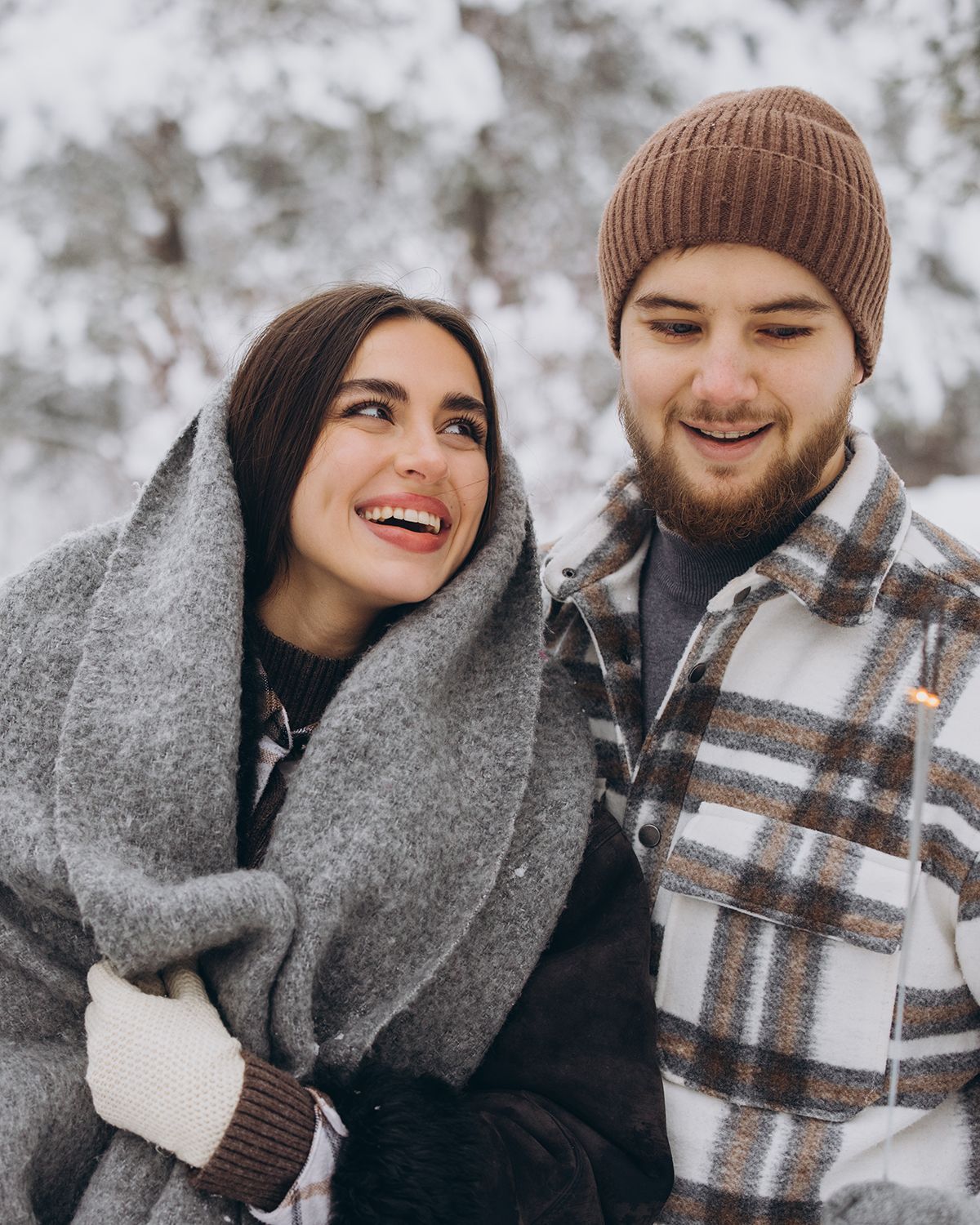 A couple smiling at each other outdoors in a snowy, wooded setting, wearing winter clothes including scarves and hats.