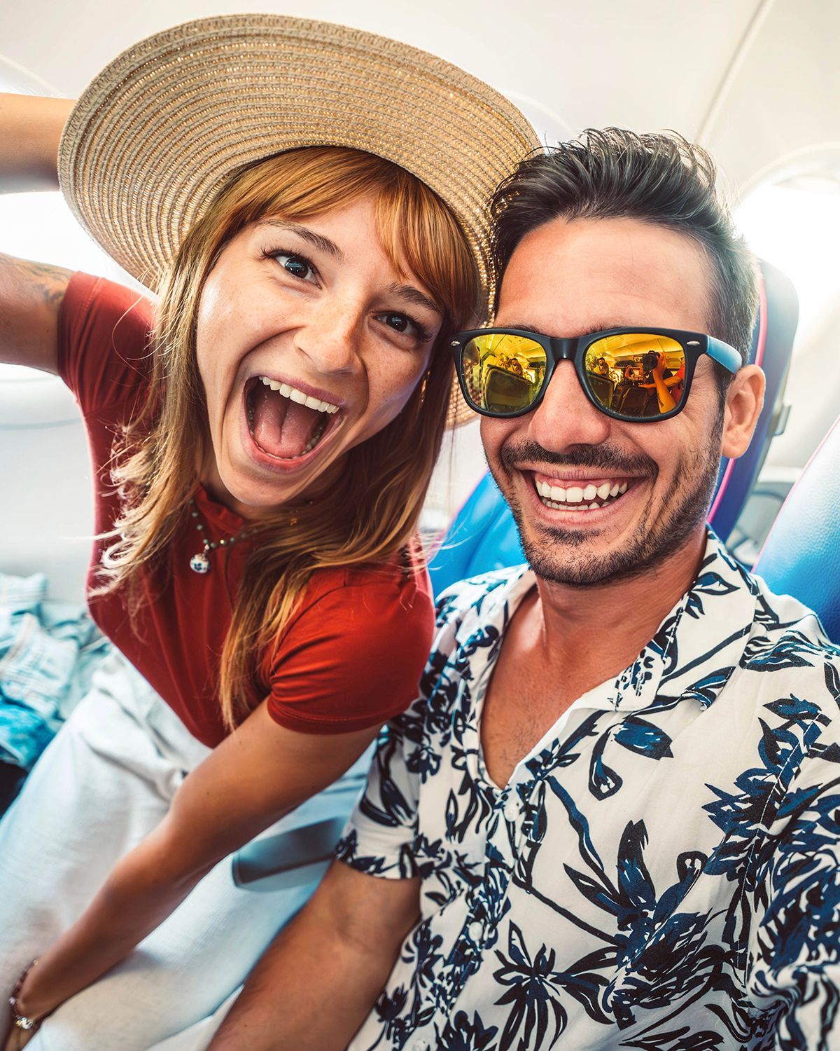 A woman wearing a straw hat and a man with sunglasses smile broadly while taking a selfie inside an airplane.