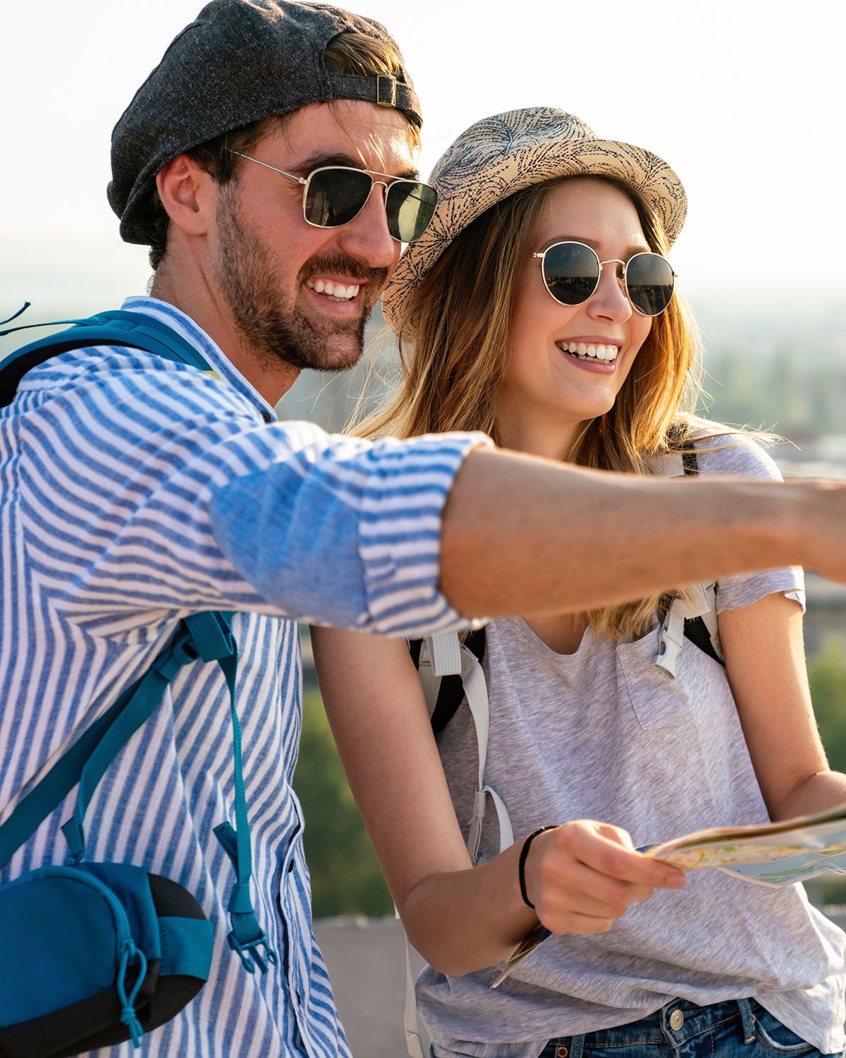 A smiling couple wearing hats and sunglasses stand outdoors, pointing at a map while on a trip.