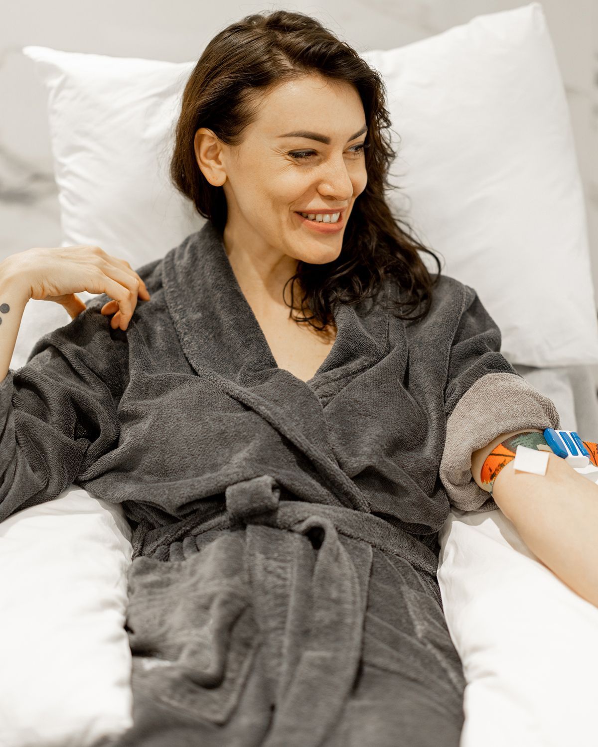 A smiling person in a gray robe resting in bed, with a medical bandage and tubes attached to their arm.
