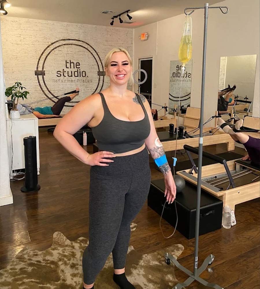 Smiling woman receiving an IV hydration drip inside a Pilates reformer studio, standing next to an IV pole.