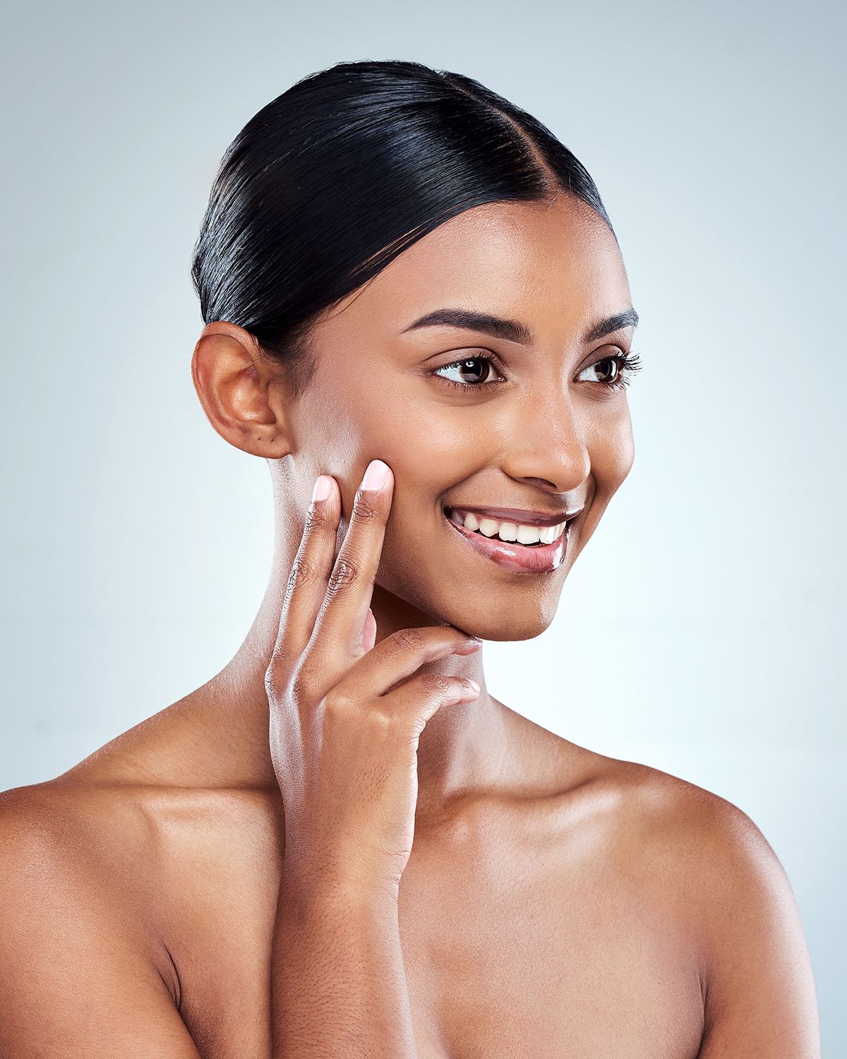 A person with smooth, glowing skin smiles while gently touching their cheek against a soft, light-blue background.