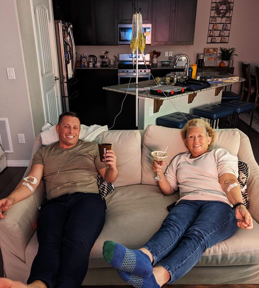 Two people on a couch receiving IVs, smiling, holding drinks in a home setting.