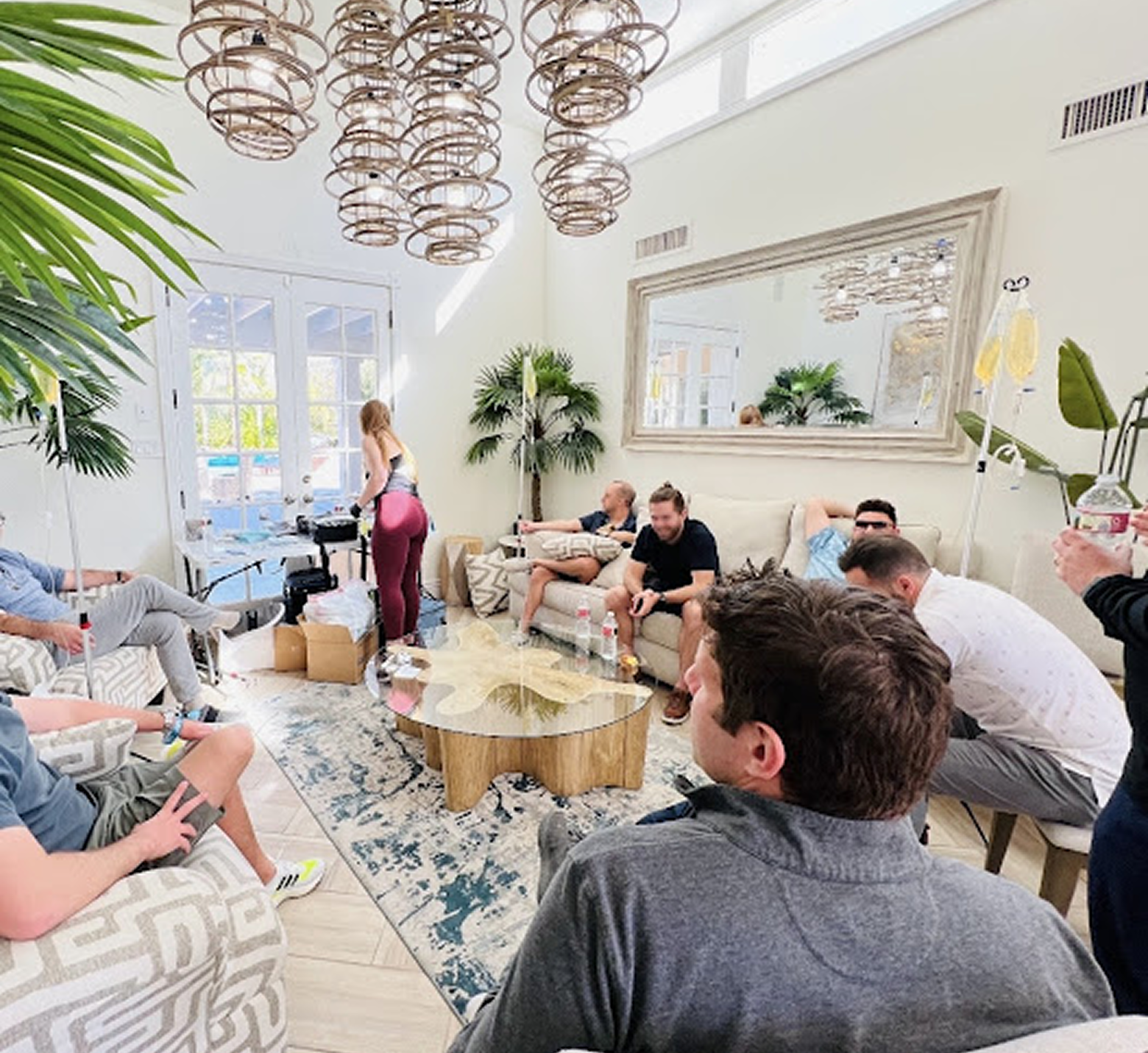A group receives IV therapy in a bright, modern living room while relaxing on couches and chairs around a coffee table.