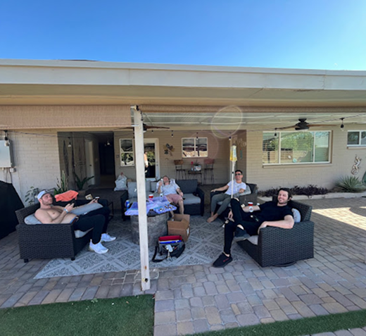 Four people relax in an outdoor patio seating area with dark wicker furniture on a paved porch under a covered roof.