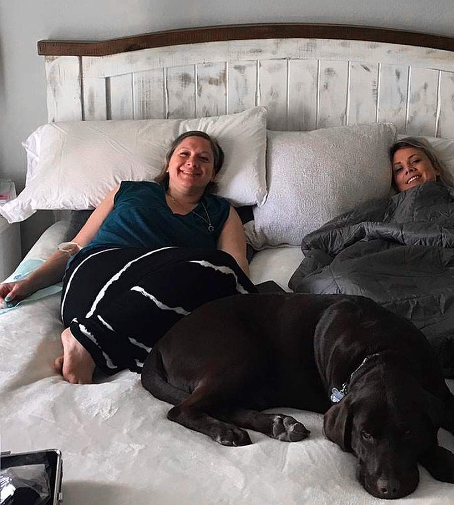 Two women and a dog on a bed. One woman smiles in a teal shirt, the other under a blanket, dark dog resting.